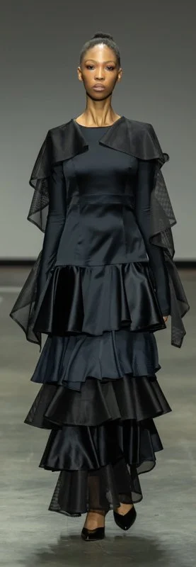 Model wearing a tiered black dress with sheer ruffled sleeves on runway.