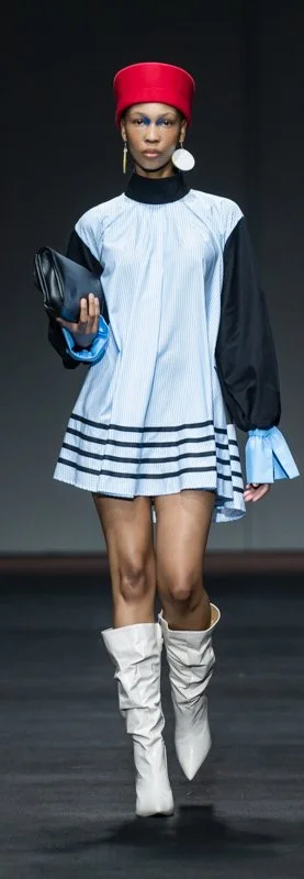 Fashion model walking down runway wearing a blue dress with black details, a red hat, knee-high white boots, and holding a black clutch.