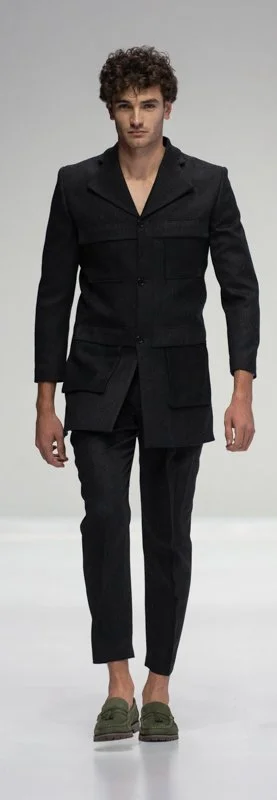 A male model wearing a black tailored suit with cropped pants and green loafers on a runway.