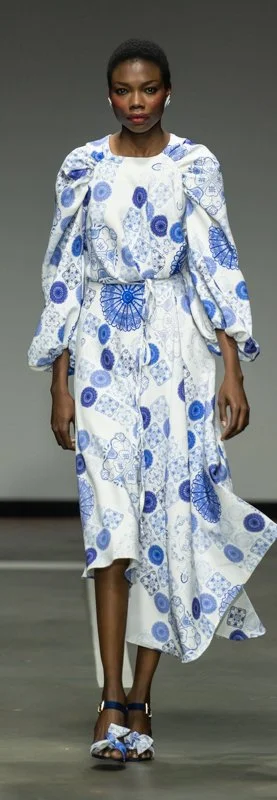 Model walking on runway wearing a white and blue patterned dress with puffy sleeves and matching shoes.