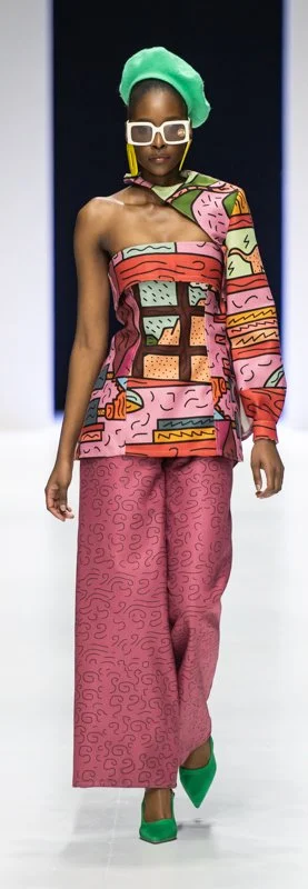Model on runway wearing colorful, patterned one-shoulder top, matching pink pants, green beret, and green heels.