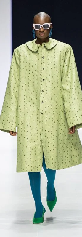 Fashion model on runway wearing green patterned coat, blue tights, green shoes, white sunglasses, and blue earrings.