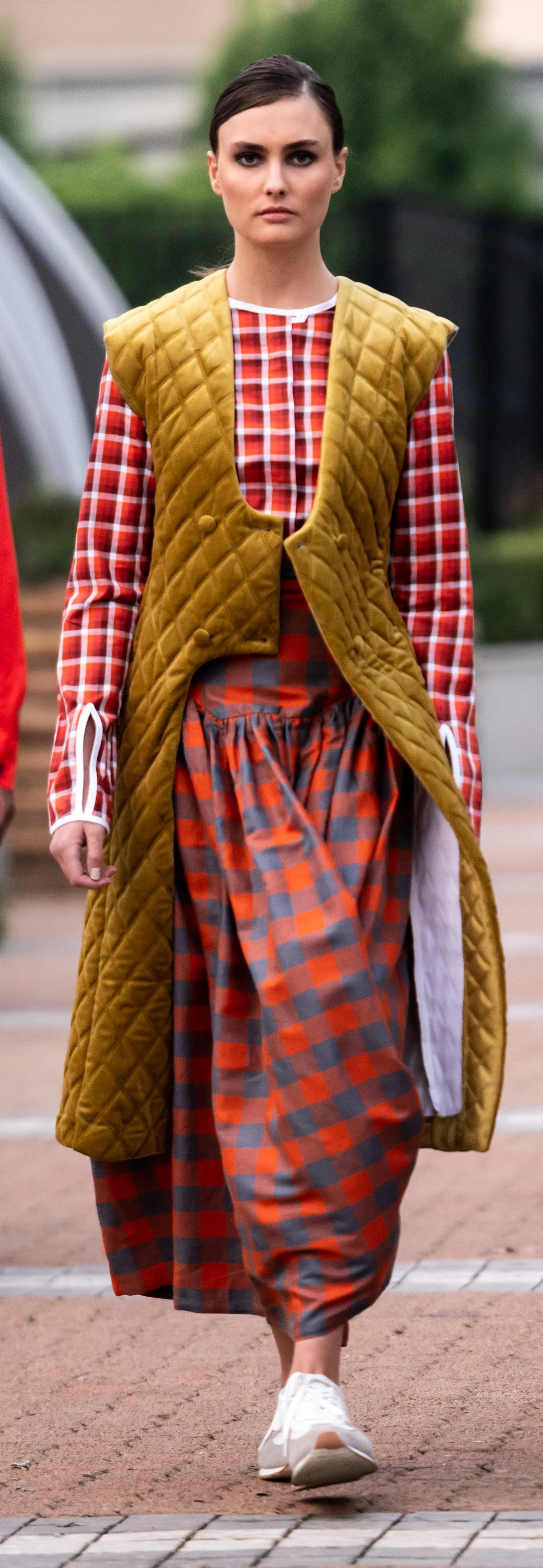 Model walking in fashion show wearing a quilted mustard yellow vest, red and white checkered shirt, red and gray checkered skirt, and white sneakers.