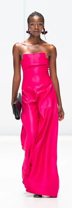 Model wearing a strapless, bright pink gown on a runway, holding a black clutch.