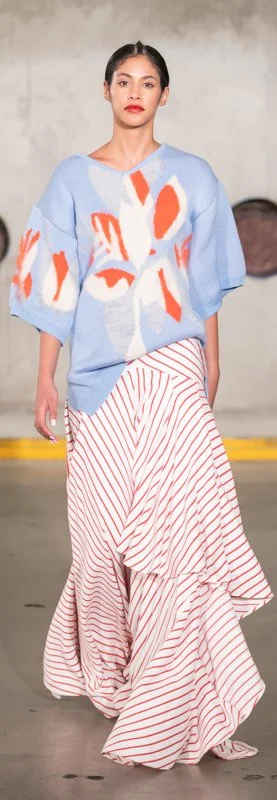 Model wearing a blue sweater with abstract red and white designs and a red-and-white striped skirt on a runway.