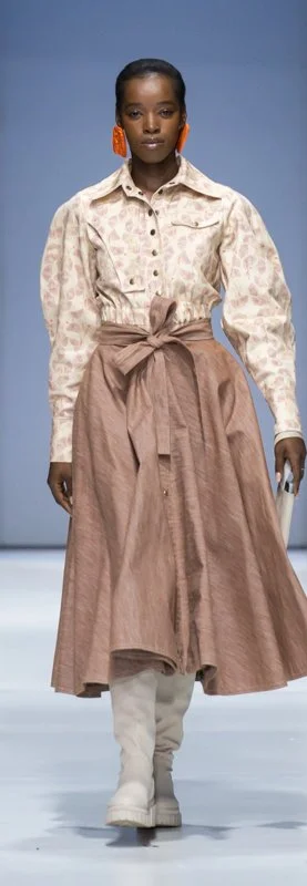 A model in a runway show wearing a floral print blouse and a long brown skirt with a tied belt, paired with white boots.