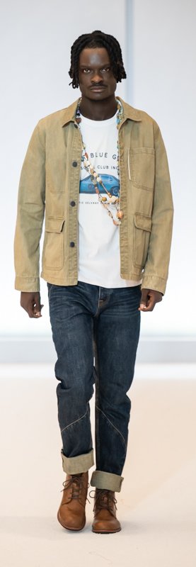 Model wearing a casual outfit with a tan jacket, graphic t-shirt, dark jeans, and brown boots on a runway.