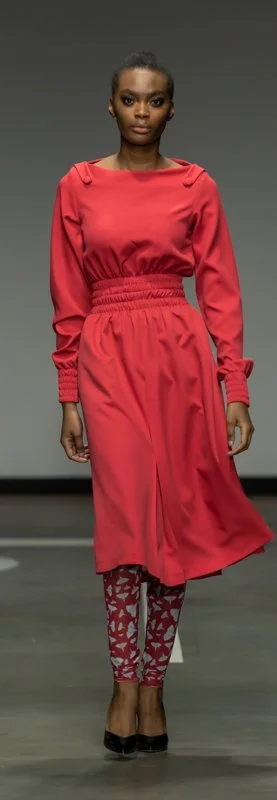 Model wearing a red dress with long sleeves and an elastic waist, paired with red patterned leggings and black heels, walking on a runway.