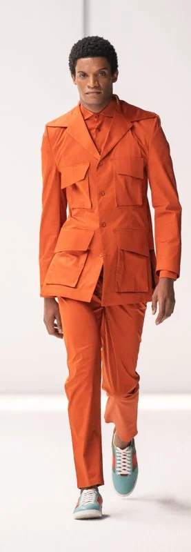 Model walking in bright orange tailored suit with multiple pockets, featuring matching pants and colorful sneakers on a white runway.