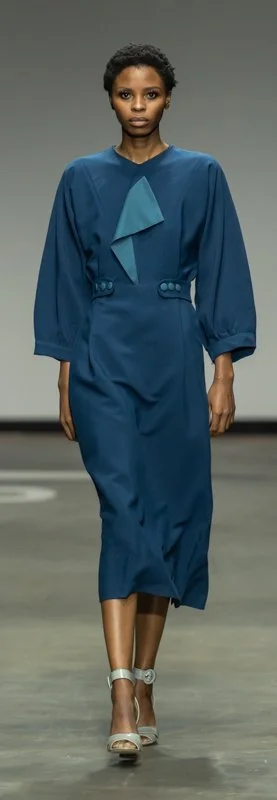 A model walks the runway in a teal-blue midi dress with long sleeves, a V-shaped neckline, button details, and wearing silver strappy heels.