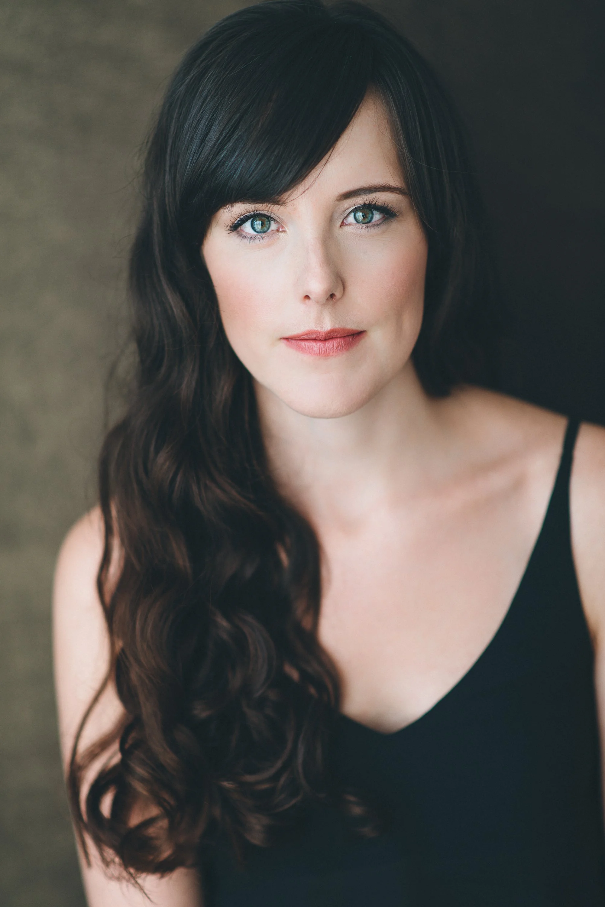Close-up portrait of a woman with long, dark wavy hair, blue eyes, and fair skin, wearing a black top with thin straps.