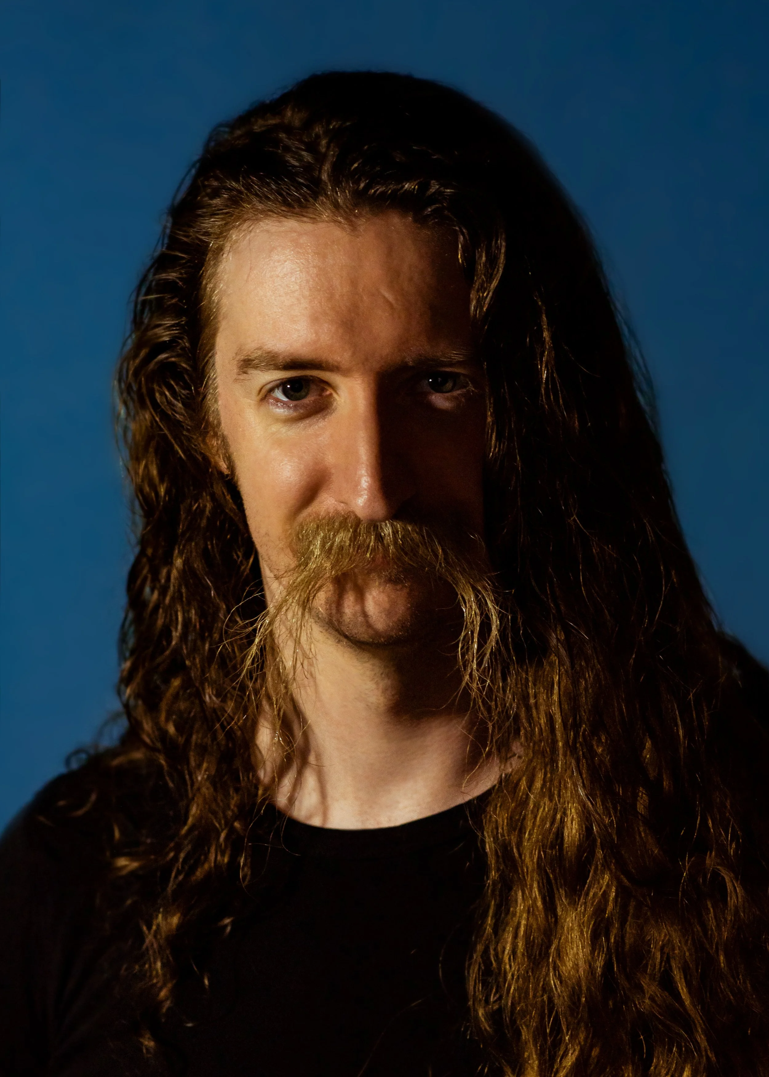 A man with long, wavy hair and a mustache, looking directly at the camera with a blue background.