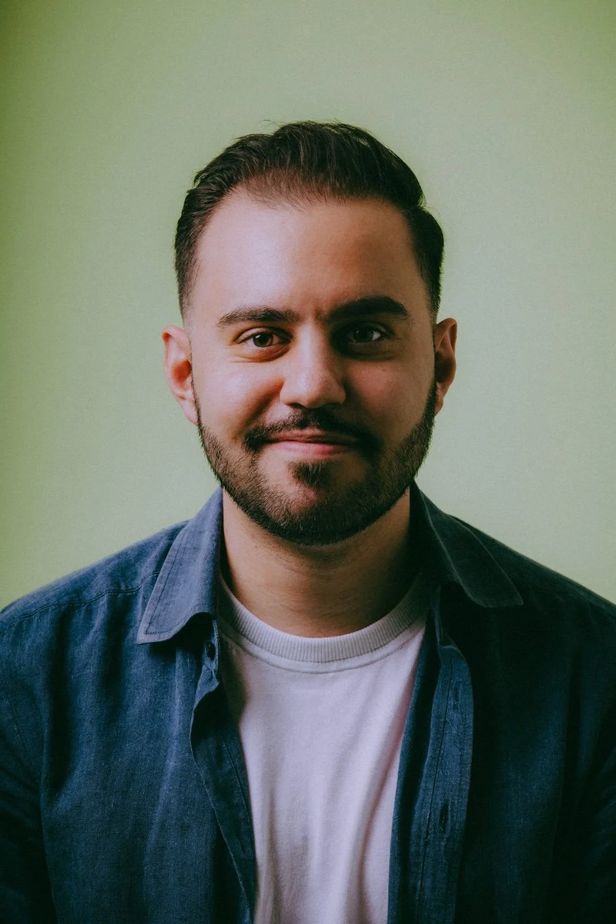 A young man with dark hair and facial hair, wearing a white T-shirt and a blue denim shirt, smiling slightly, with a light green background.