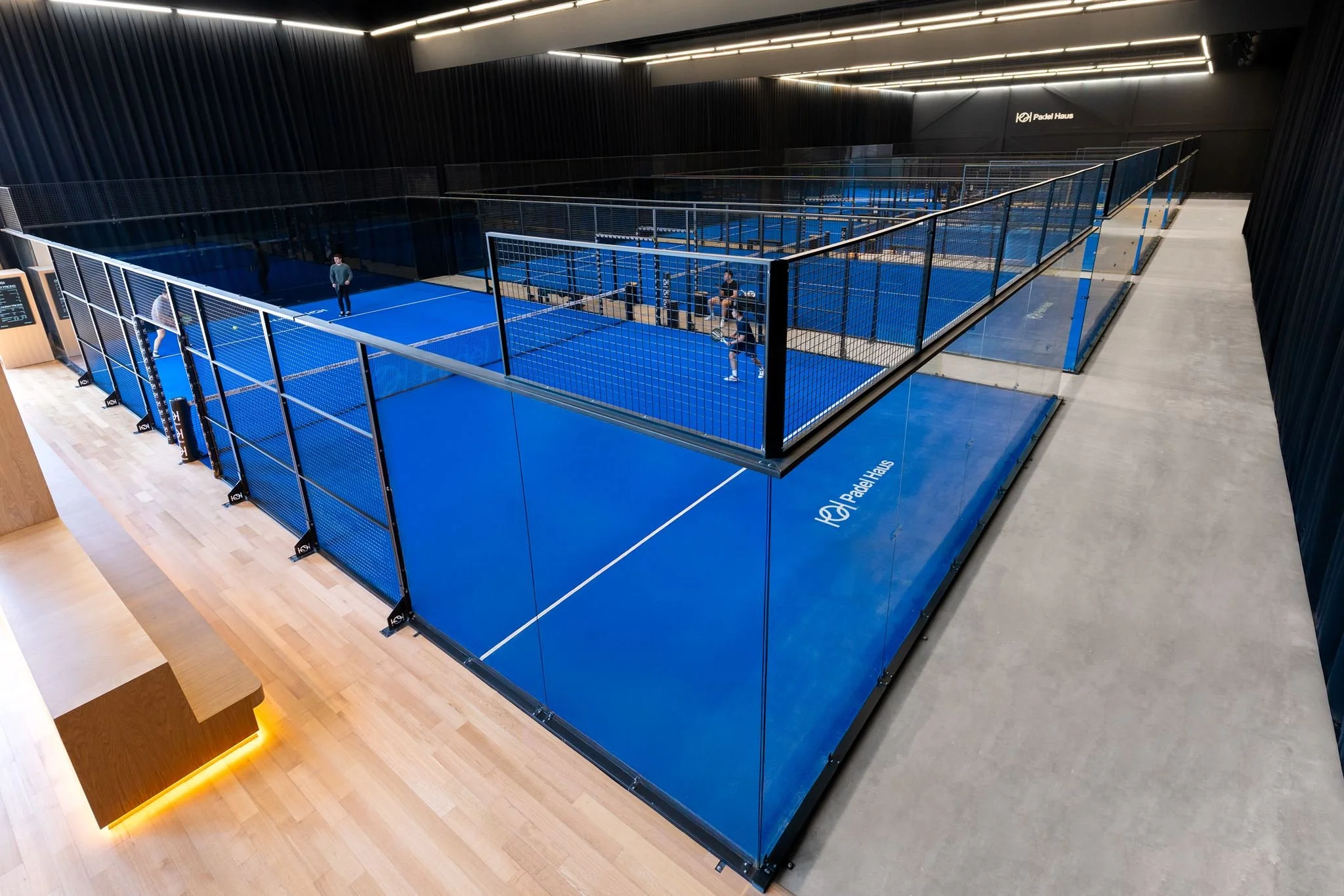 Indoor pickleball court with blue flooring, enclosed by fencing, and players practicing, situated in a modern gym with black walls and ceiling.