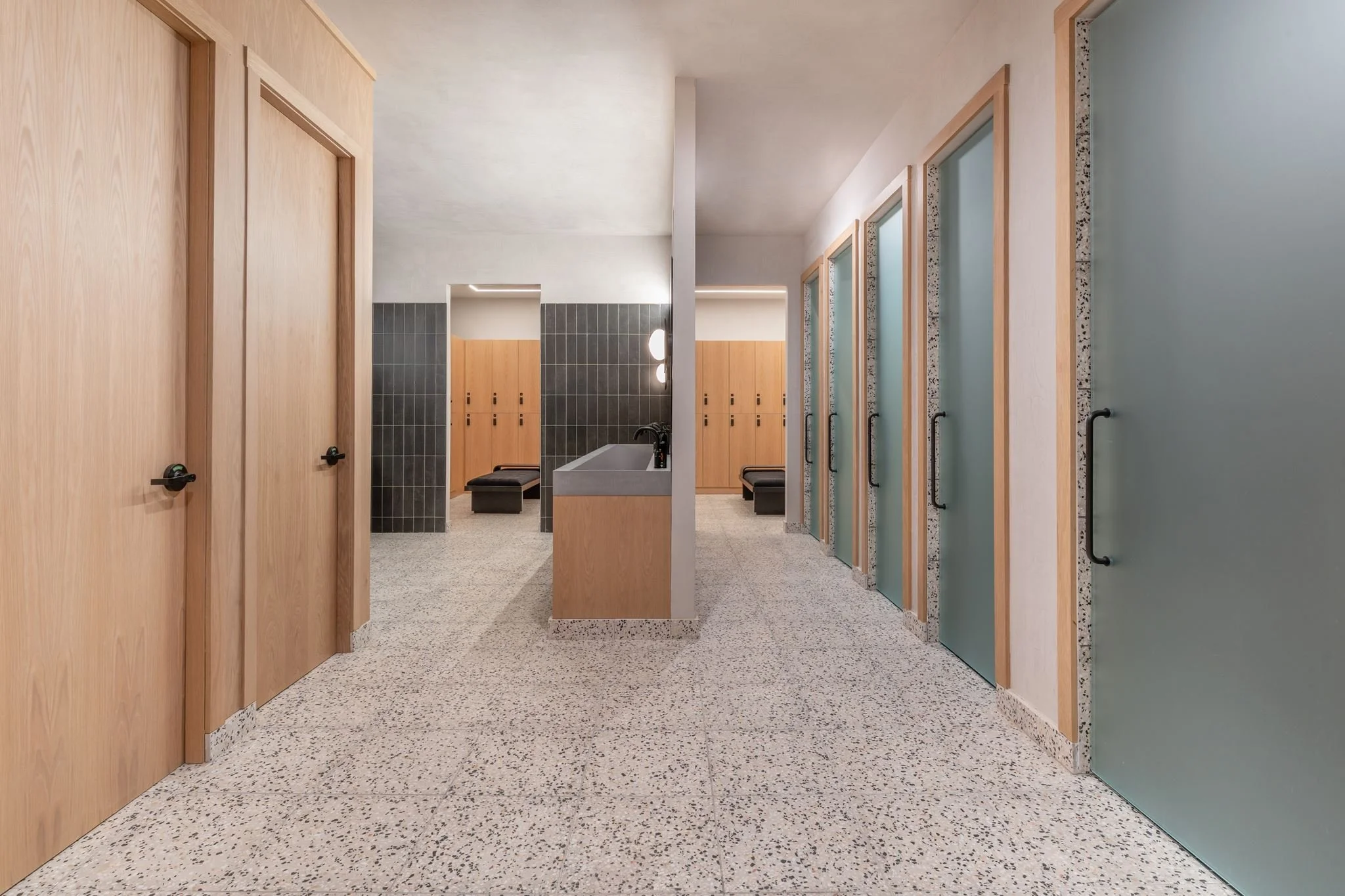 Modern changing room or locker room with wooden doors on the left, a sink in the center, and lockers on the right, with a terrazzo floor and minimalist decor.