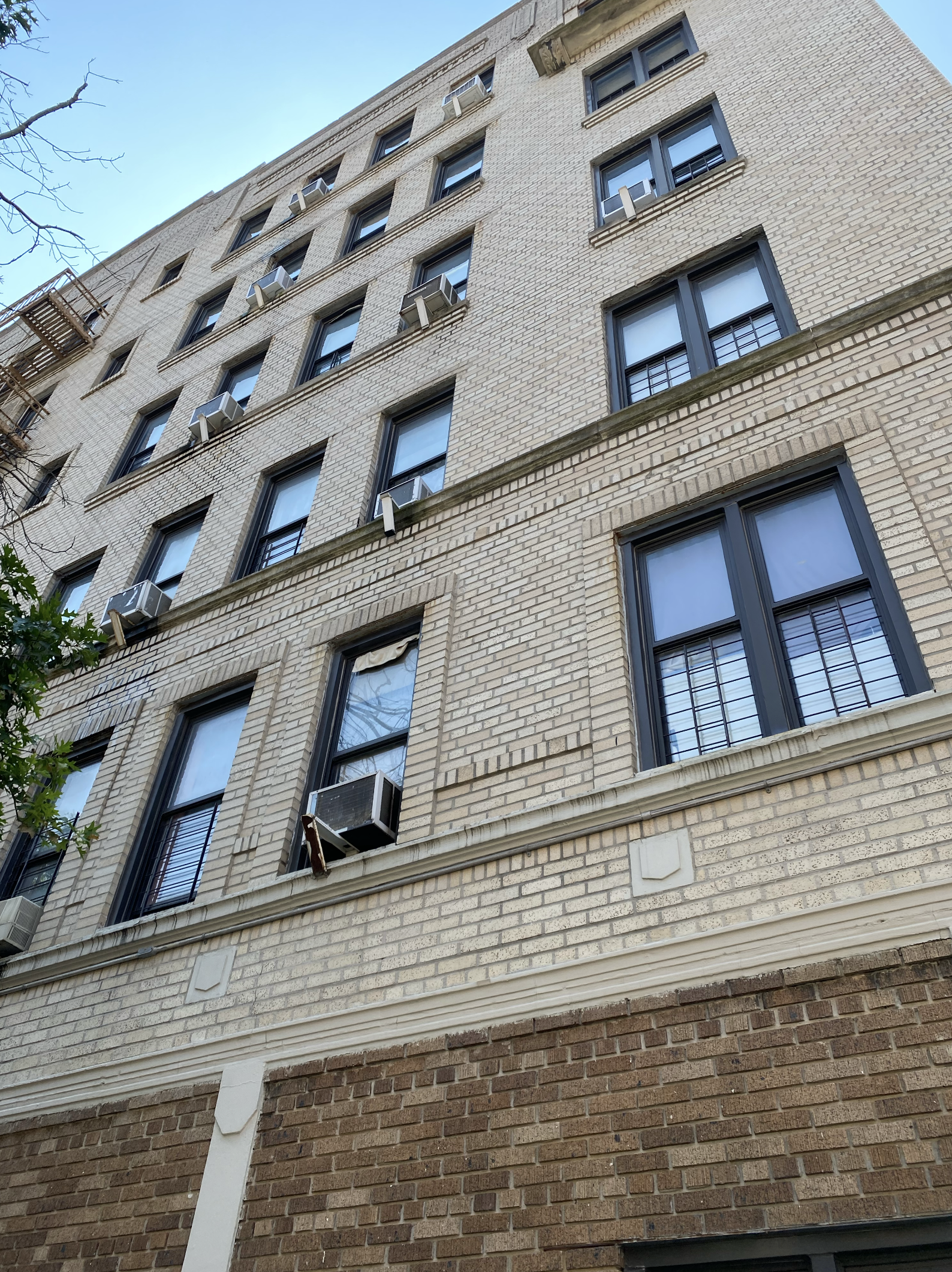 View of a multi-story brick building with several windows fitted with air conditioning units, some with blinds or curtains, and a fire escape on the side, under a clear blue sky.