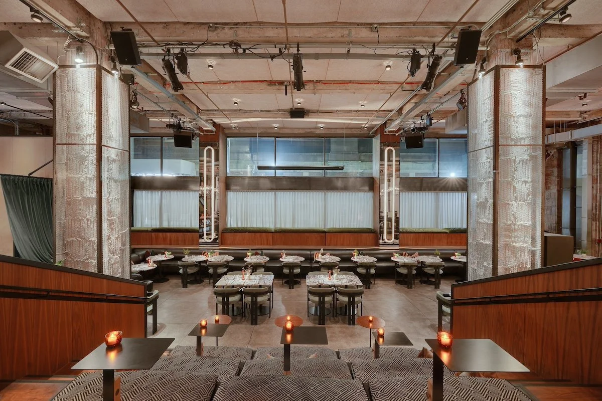 Interior of a modern restaurant or lounge with a stage, tables with flower centerpieces, cushioned window seats, and dim candle lighting.