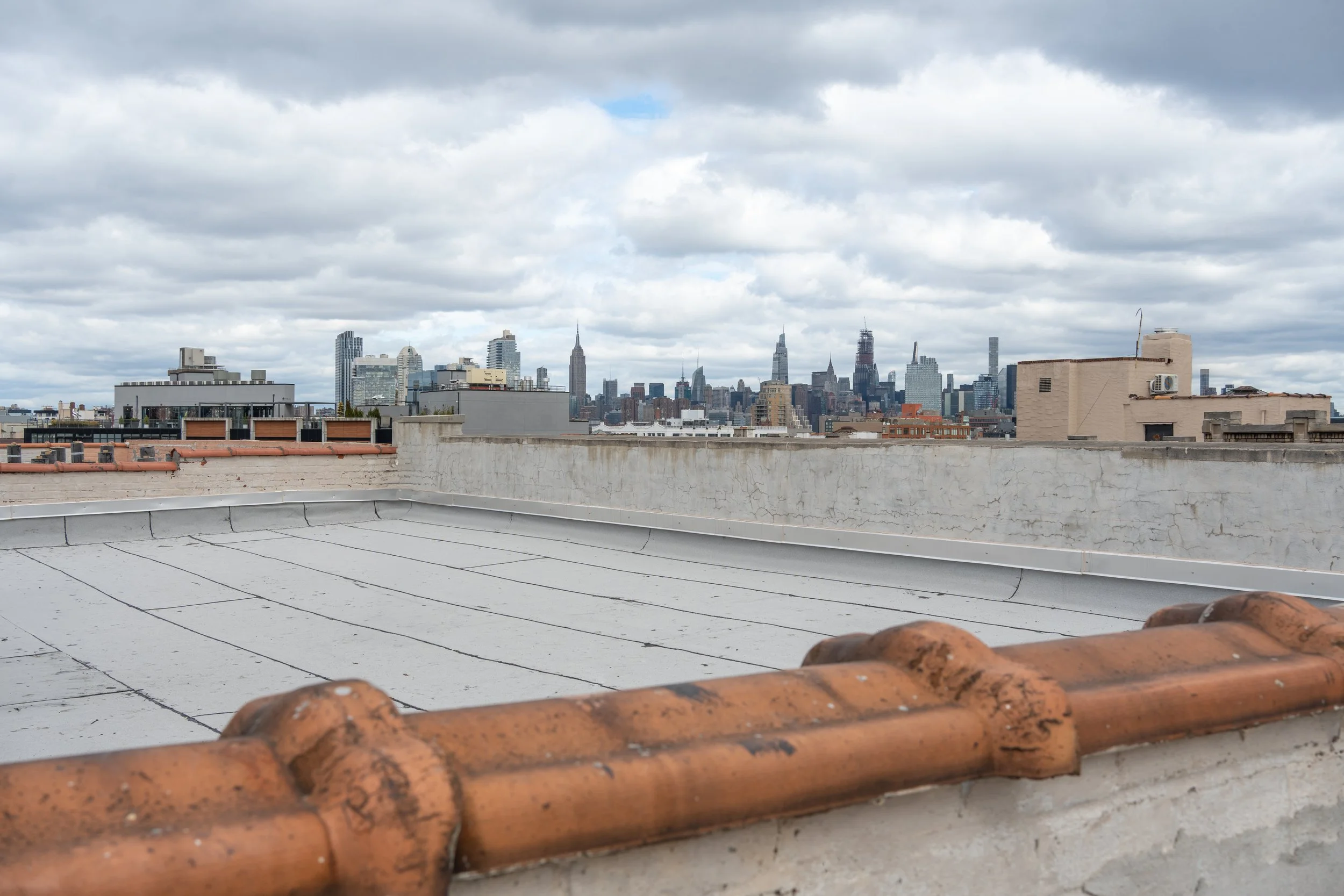 City skyline with tall buildings and skyscrapers under a cloudy sky, viewed from a rooftop with reddish-brown roof tiles and a white concrete parapet.