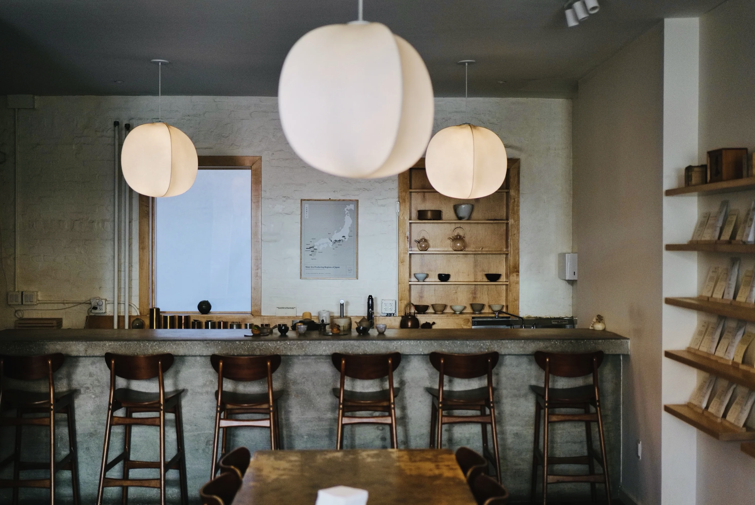 A cozy coffee shop or cafe interior with hanging pendant lights, a concrete counter with wooden bar stools, and a rustic wooden shelf with bowls and decorative items on the white brick wall.