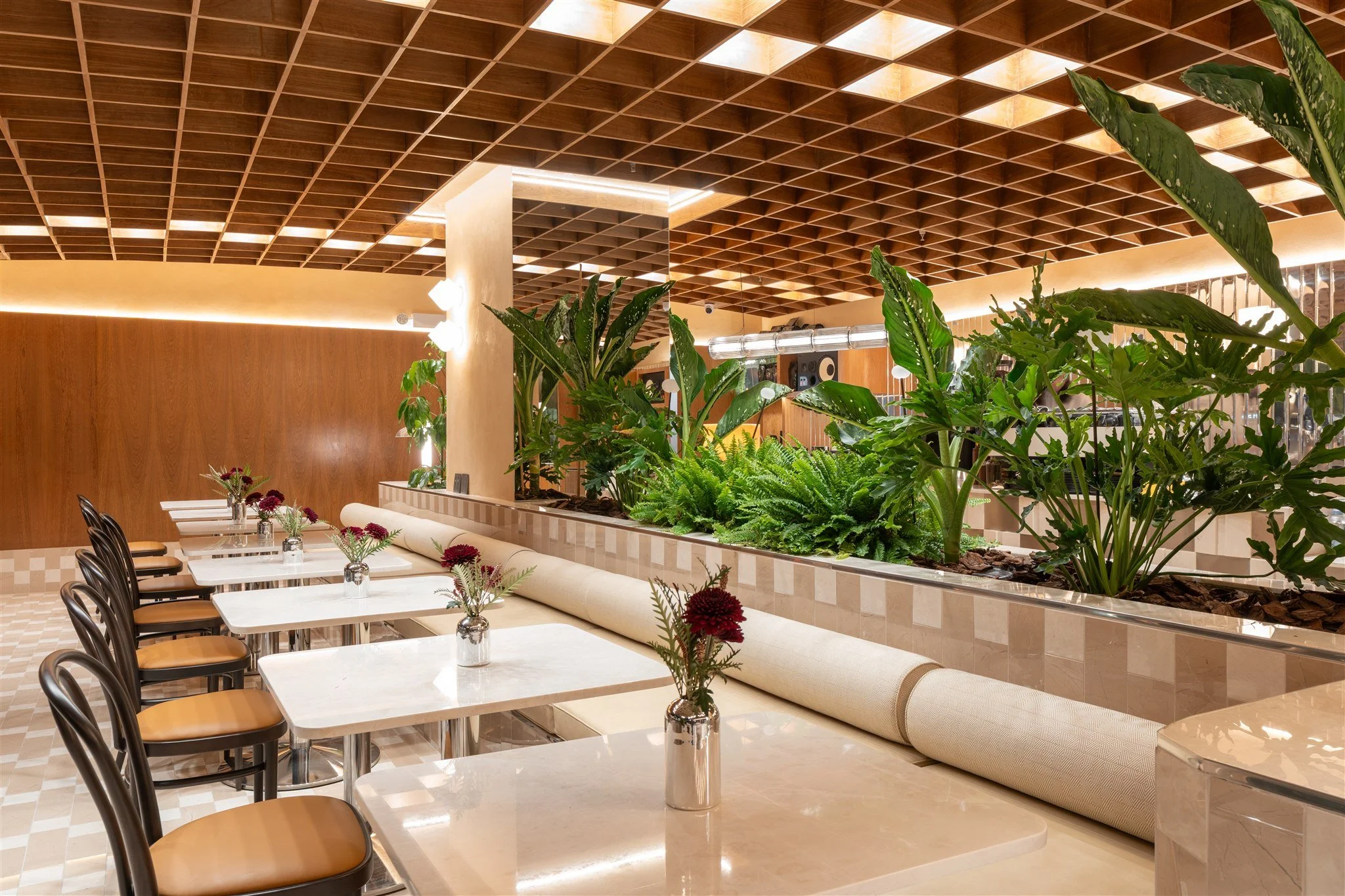 Interior of a modern restaurant with white tables, brown chairs, and decorative vases with flowers. There is a raised planter with large green plants in the middle of the space, a textured wood ceiling, and soft warm lighting.