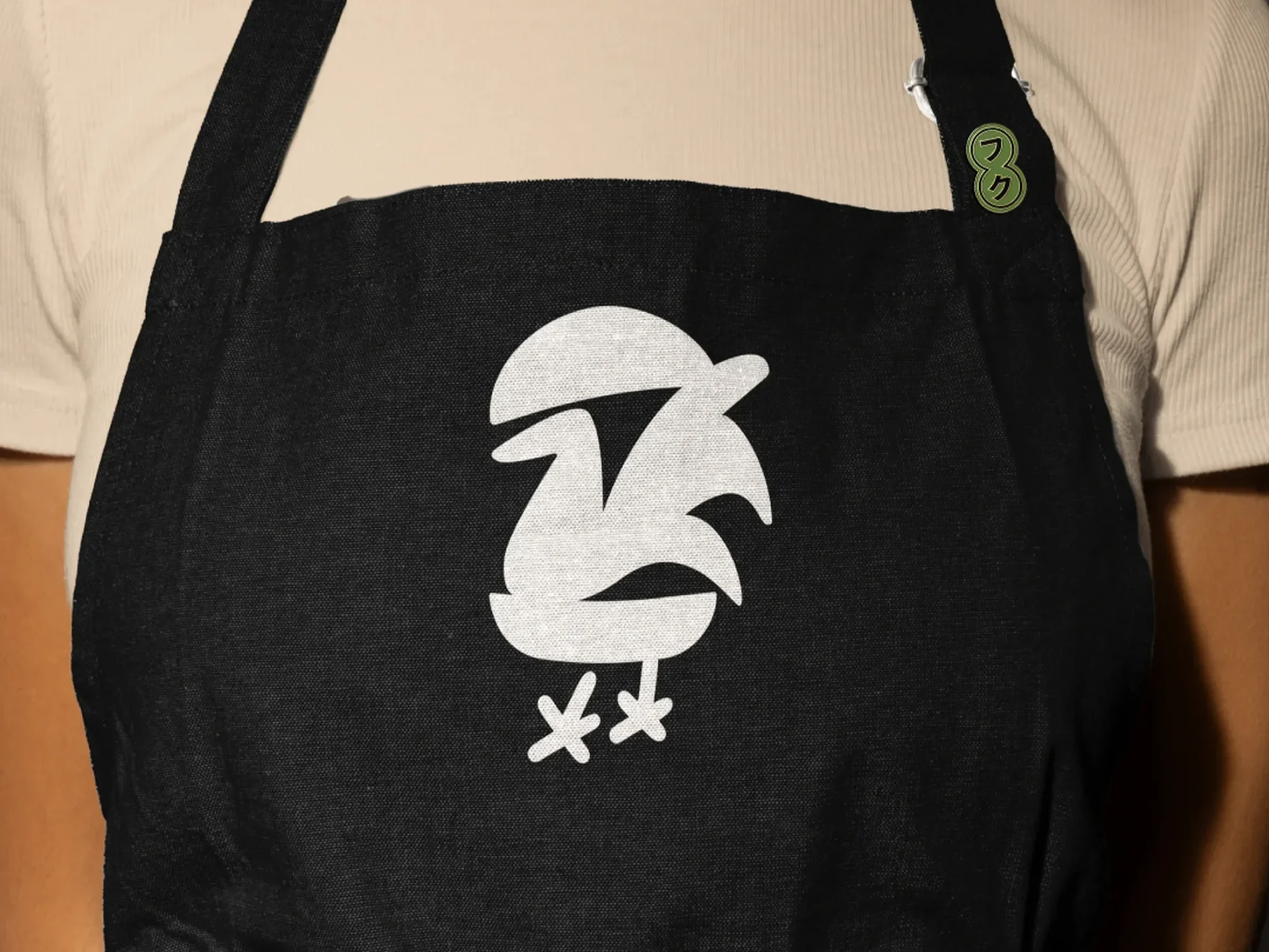 A person wearing a beige shirt and a black apron with a white rooster design on it.