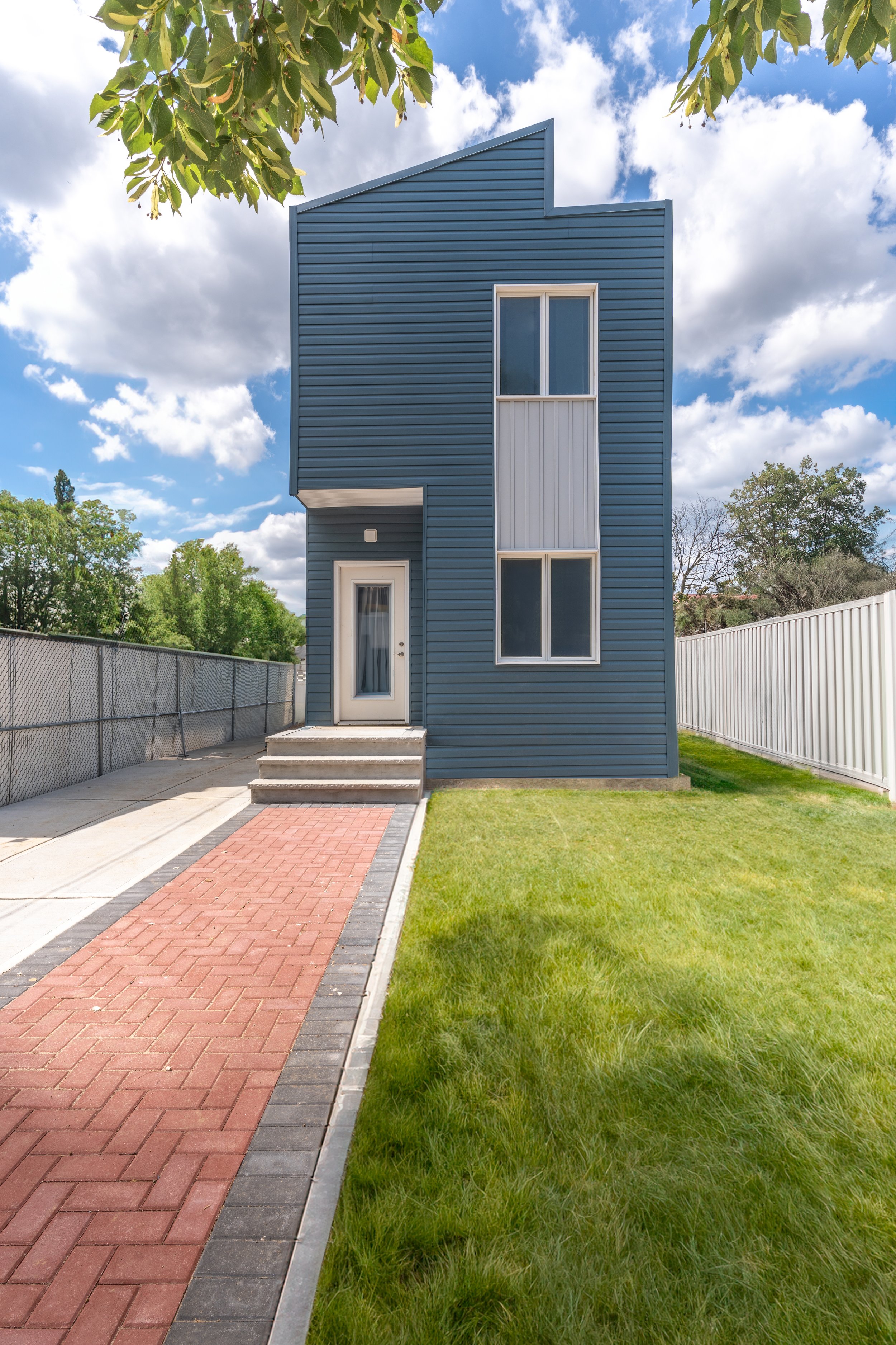 Modern blue house with white trim, two large upper windows and a front door with steps, a red brick pathway, green lawn, and a white fence, under a partly cloudy sky.