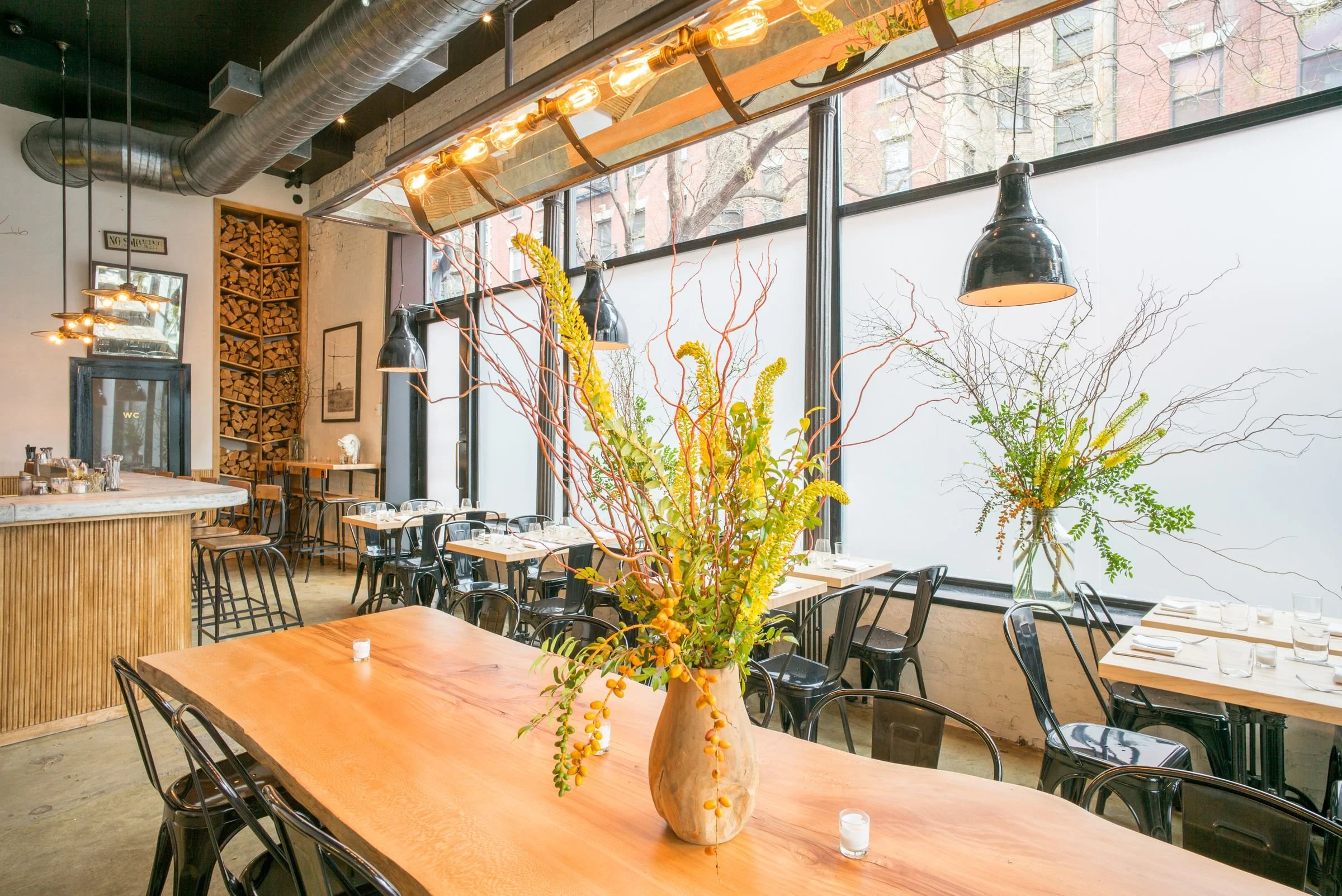 The interior of a restaurant with large windows, a central floral arrangement with yellow flowers and green foliage in a large beige vase, black chairs around wooden tables set for dining, pendant lights with black shades, and an exposed ceiling with large silver ductwork.