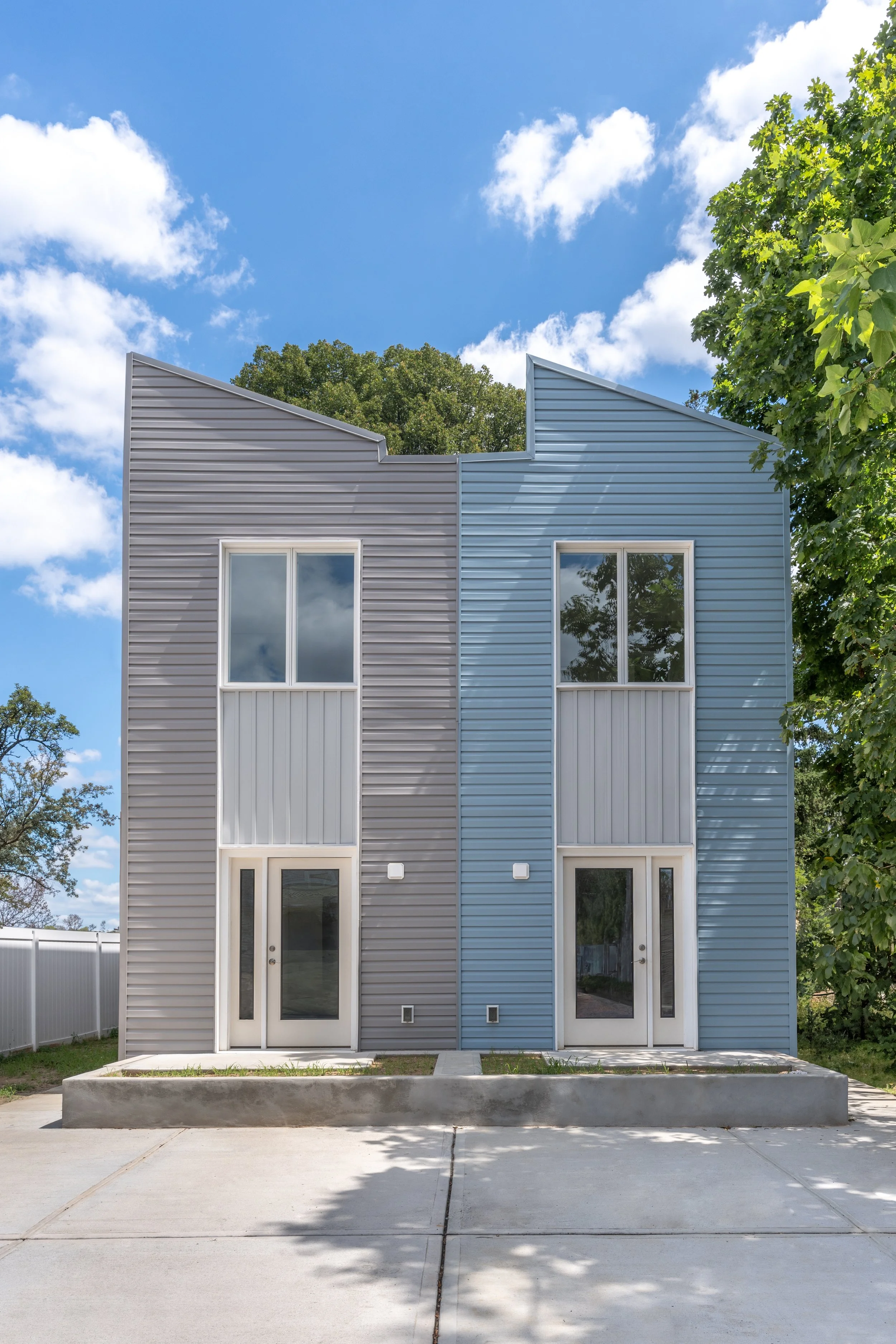 Modern duplex with gray and blue metal siding, large windows, and concrete walkway in front, surrounded by green trees under a partly cloudy sky.