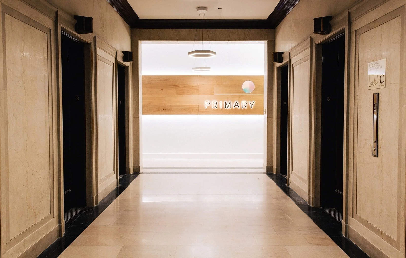 Lobby area with elevators on either side, wood-paneled wall with the word 'PRIMARY' illuminated, and a modern light fixture hanging from the ceiling.