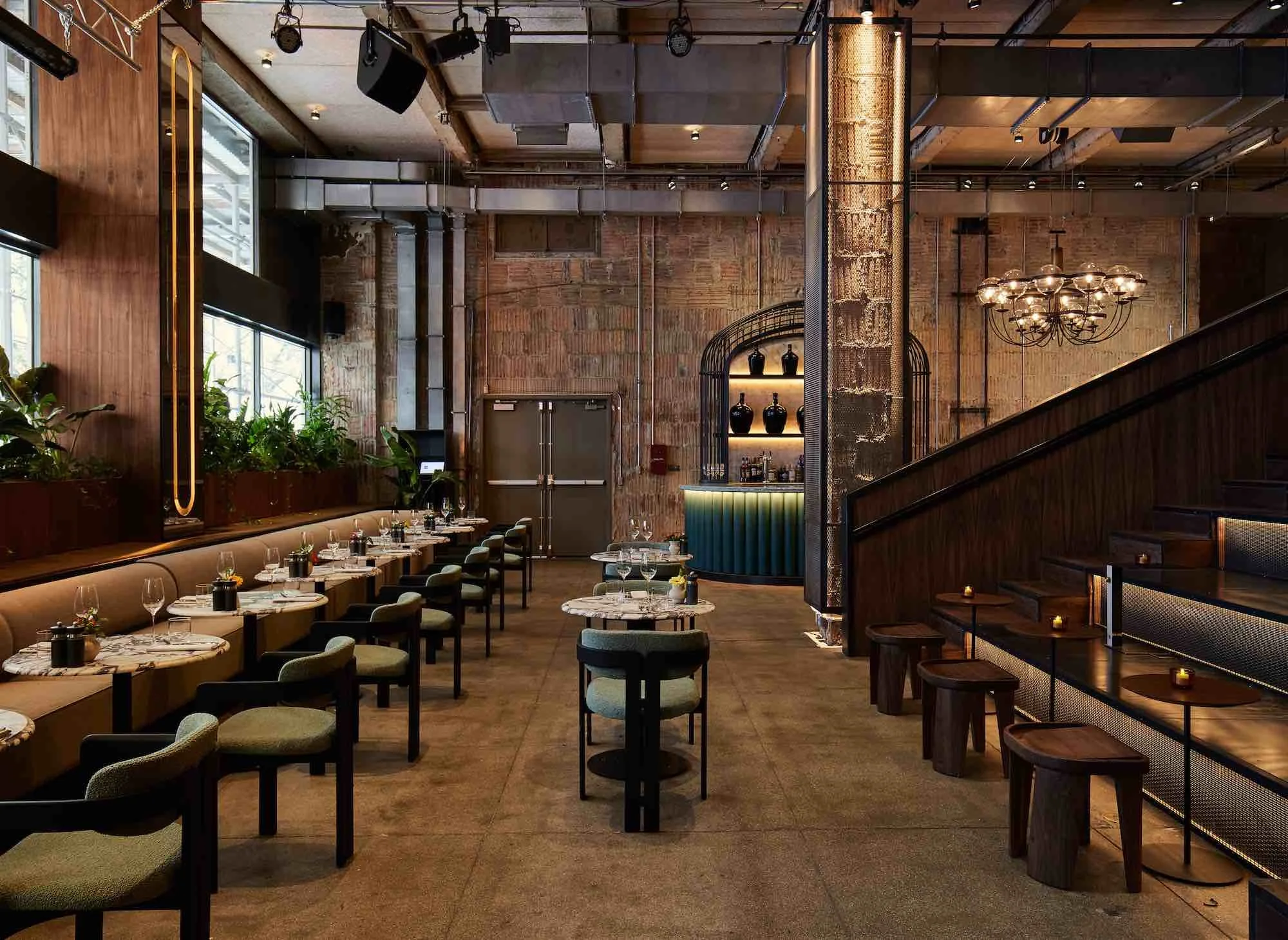 Interior of a modern restaurant with wood and metal decor, chandeliers, seating with green and beige chairs, tables with wine glasses, and large windows letting in natural light.