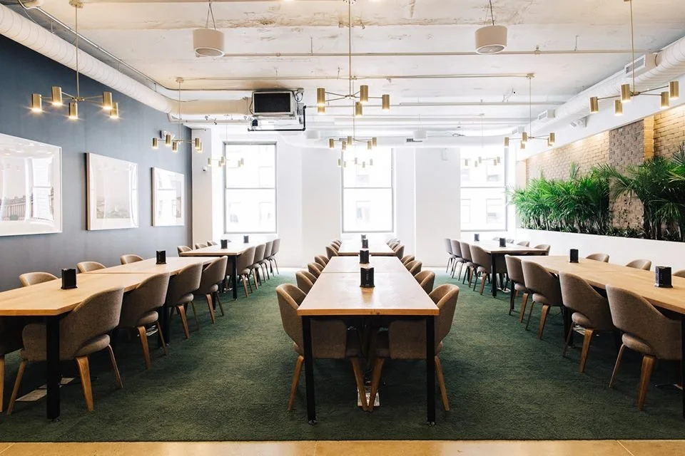 A modern conference room with long wooden tables, beige chairs, large windows, green plants, and minimalistic decor.