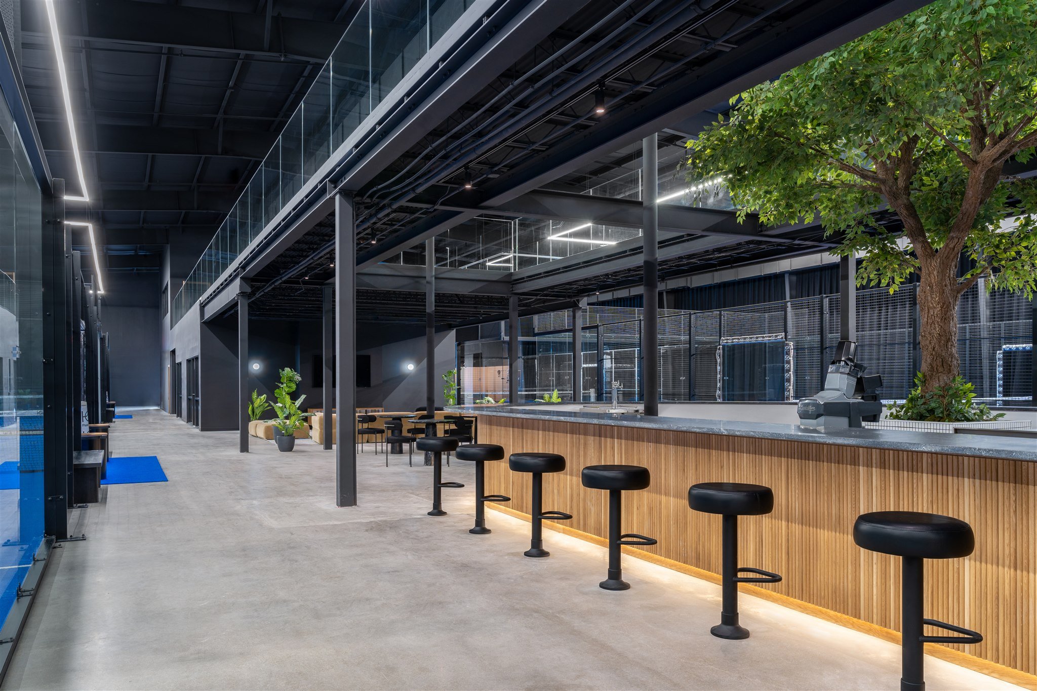 Modern indoor public space with a bar counter, black stools, indoor greenery, and a large tree, featuring black metal and glass architecture.