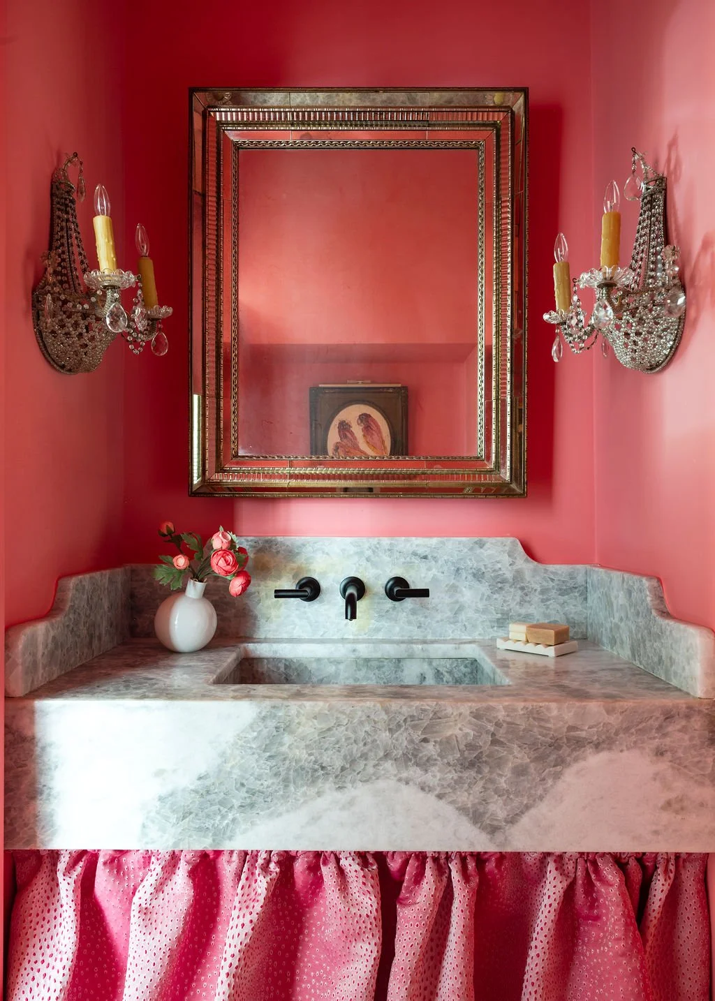 A vanity with a marble countertop, a large rectangular ornate mirror, pink walls, two crystal wall sconces with candles, a small vase with pink flowers, and a soap dish with soap bars.