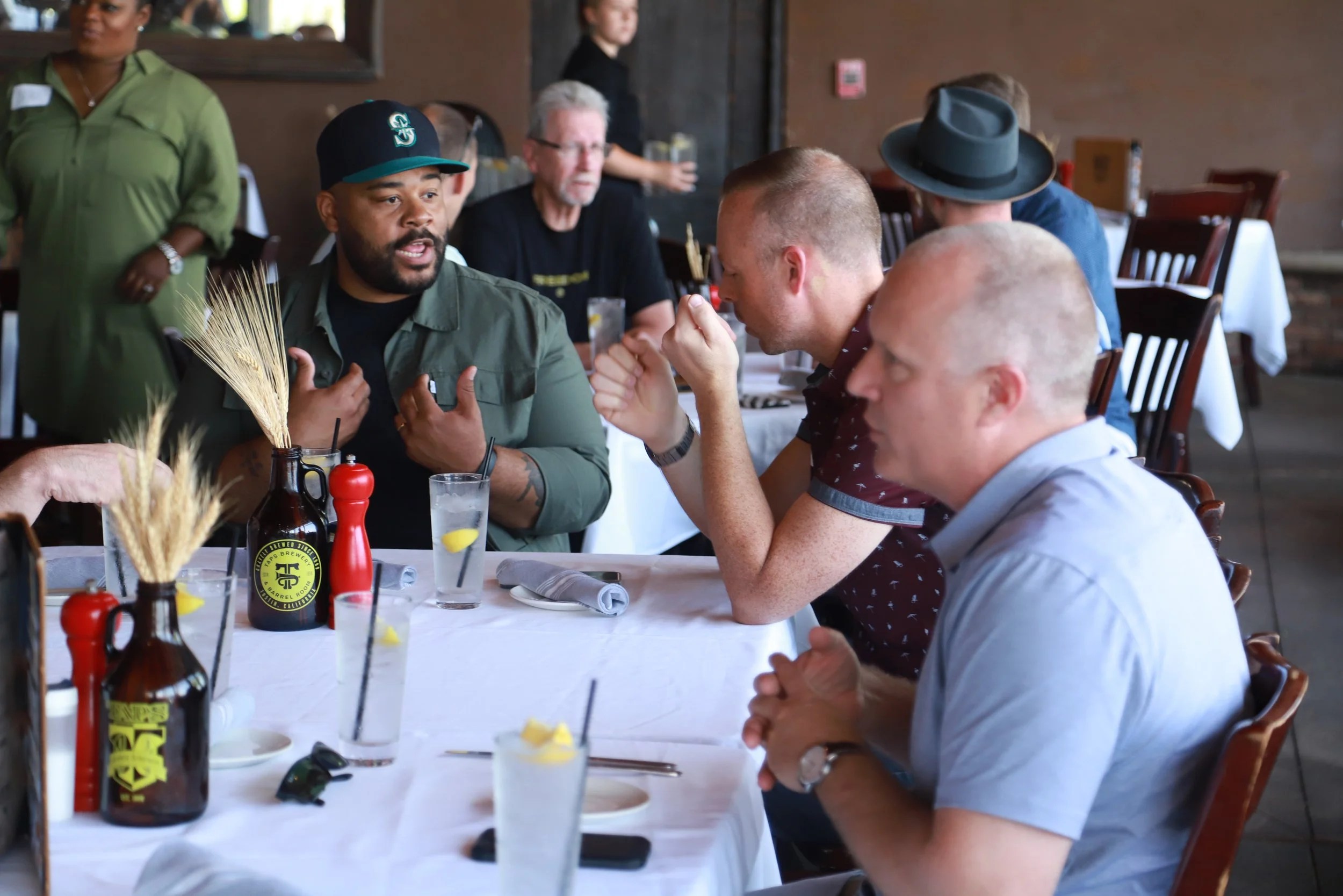 Group of people sitting at a restaurant table, engaged in conversation. Some are drinking drinks, with condiments and tableware on the table, in a casual dining setting.