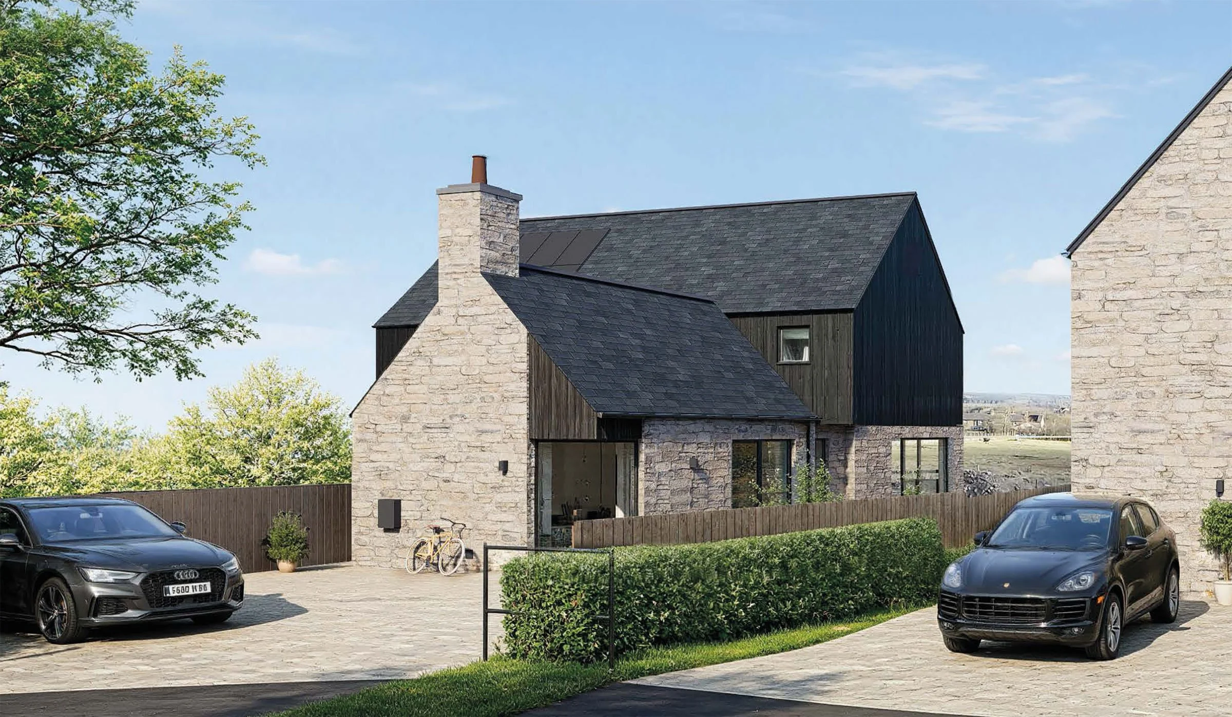 Modern house with stone and black wooden exterior, large windows, and a chimney, surrounded by a driveway with parked cars and a wooden fence, under a blue sky with some clouds.