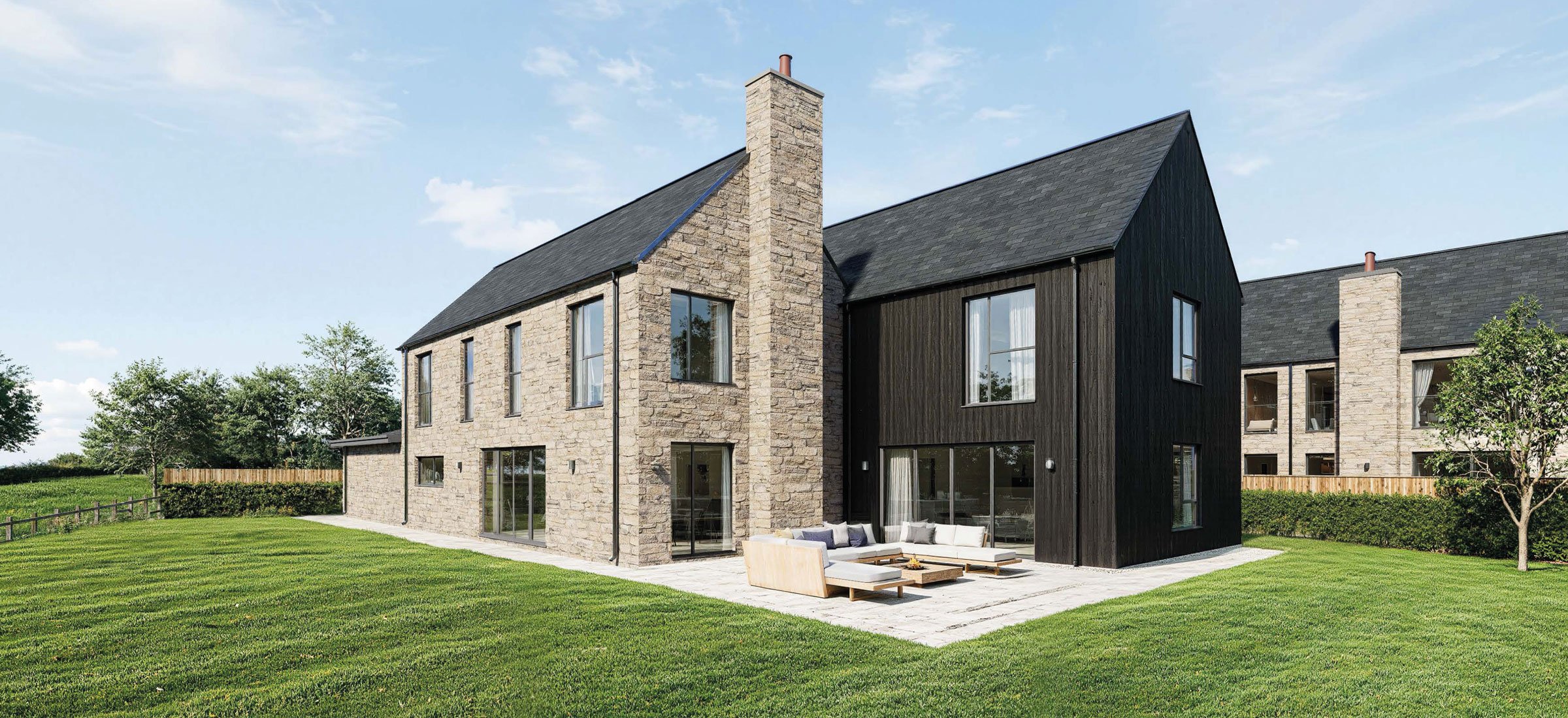 Modern residential buildings with stone and black wooden exterior, large windows, a patio with outdoor seating, green lawn, and trees under a blue sky.