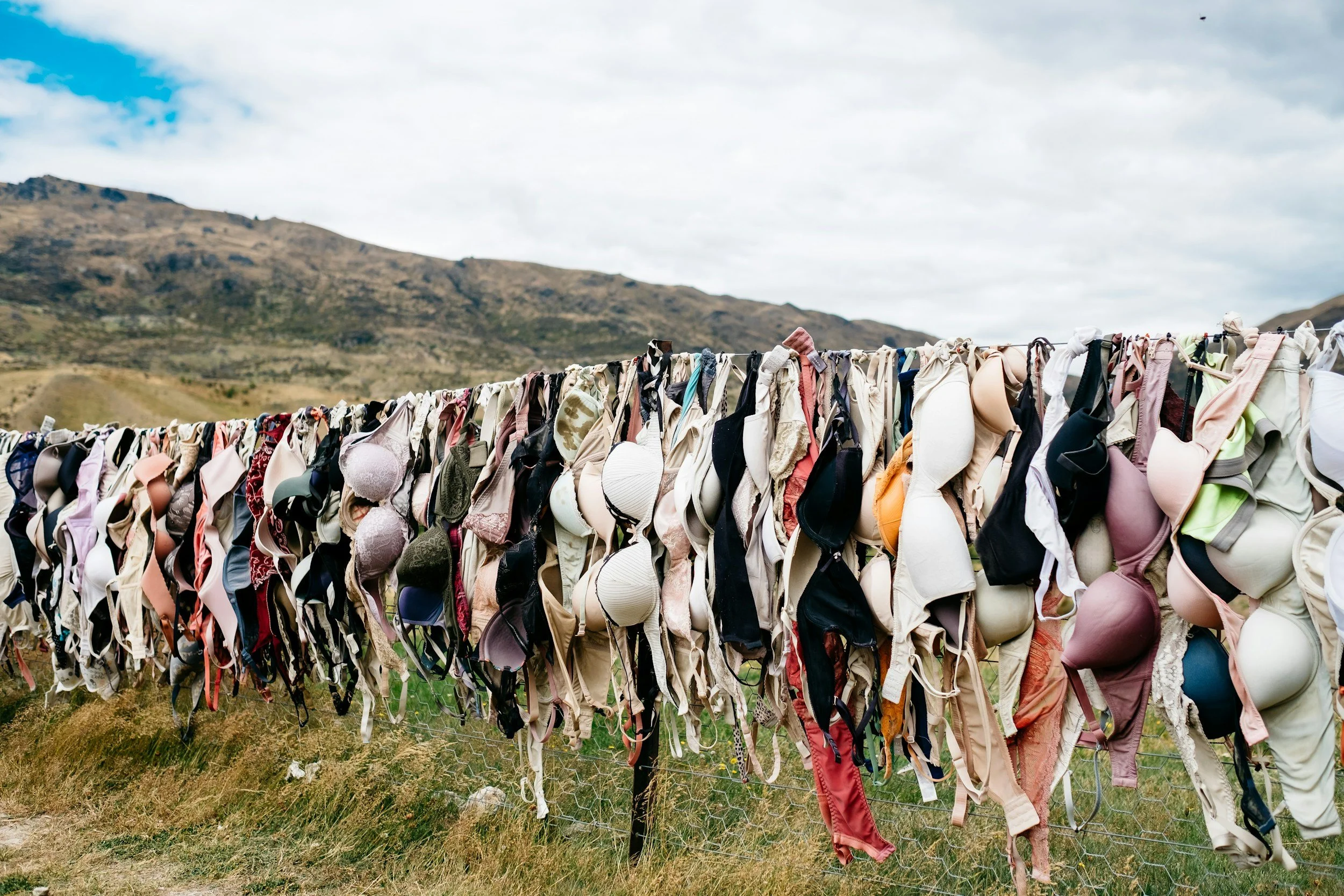 A long line of bras hanging on a makeshift clothesline outdoors with mountains and cloudy sky in the background.