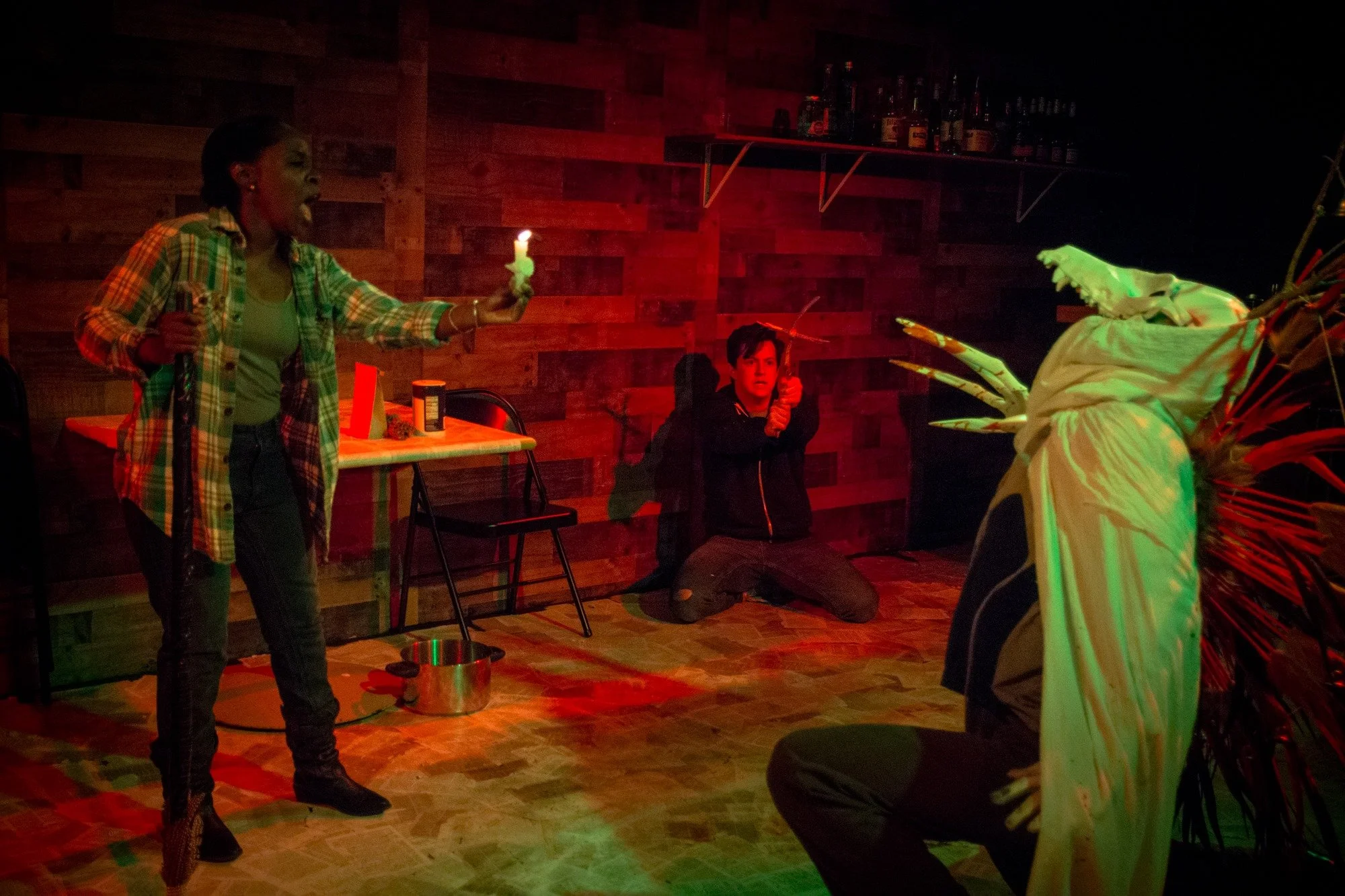 A woman with a walking stick holds a candle in the direction of a monster. A young man holds a cross in the corner.