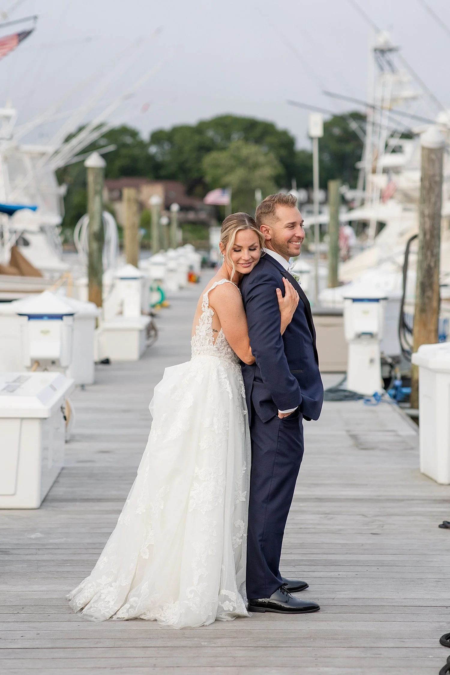  bride hugs groom from behind while standing on dock by water 