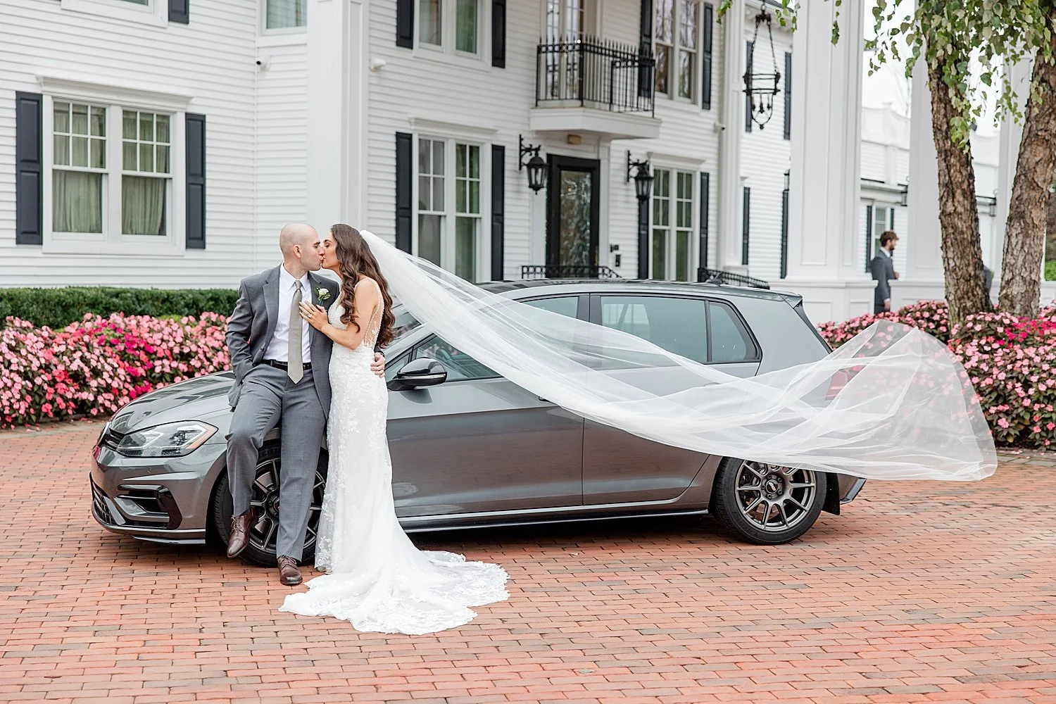  couple kiss by car with bride’s veil flowing behind her 