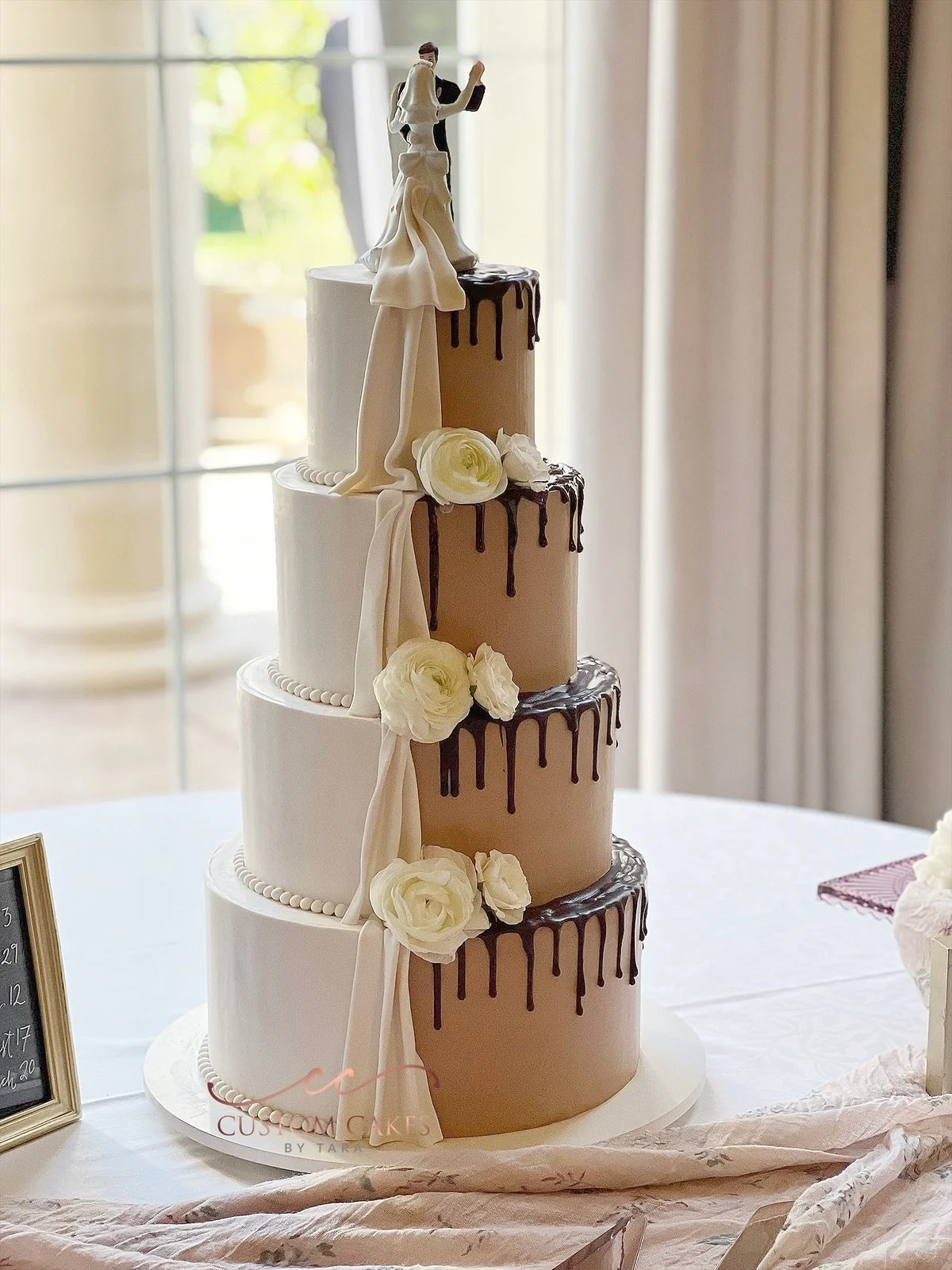 Four-tier wedding cake with white and beige frosting, decorated with white roses and brown drips, topped with a bride and groom figurine, sitting on a table near a window.