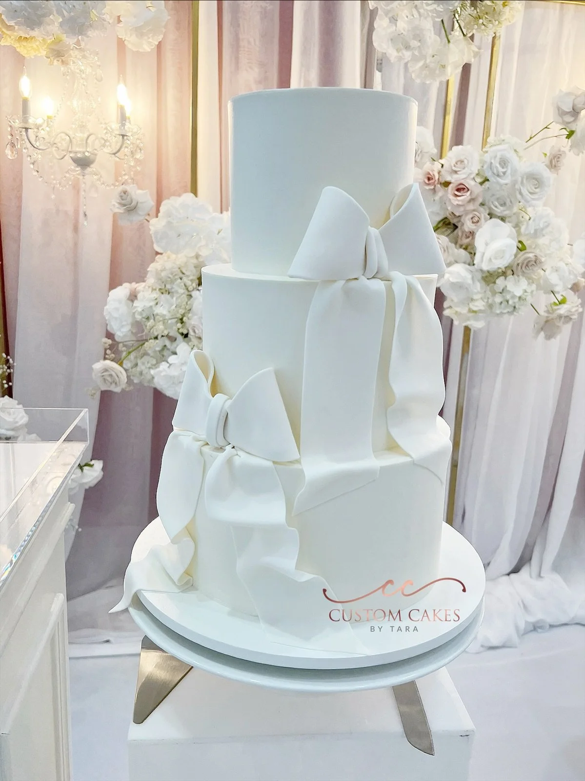 White tiered wedding cake with large fondant bows, displayed on a stand with a 'Custom Cakes by Tara' logo, surrounded by floral decorations and a chandelier in the background.