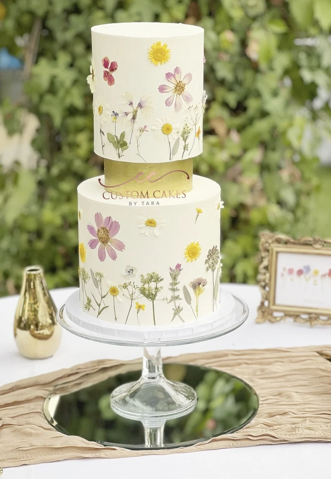 Two-tiered wedding cake decorated with pressed flowers, displayed on a glass cake stand with a mirror base, set outdoors with greenery in the background and framed sign nearby.