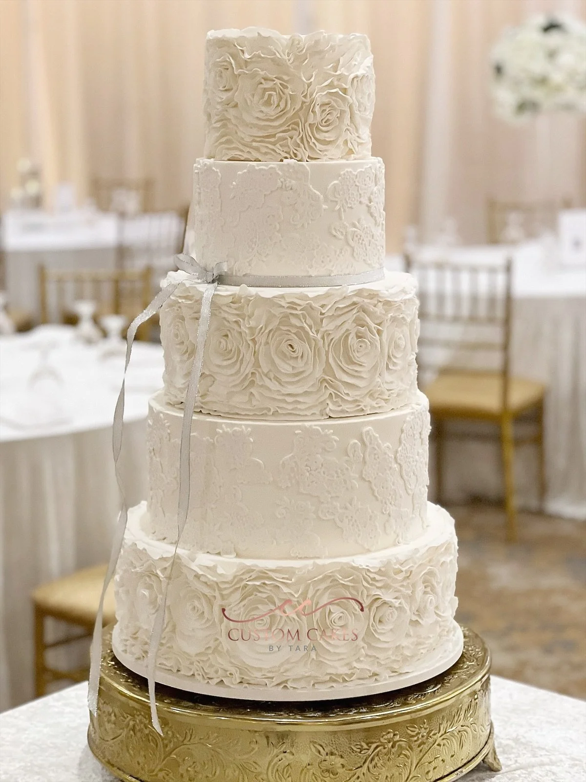 A tall, multi-tiered white wedding cake decorated with intricate piping and fondant roses, placed on a gold stand in a decorated event hall.
