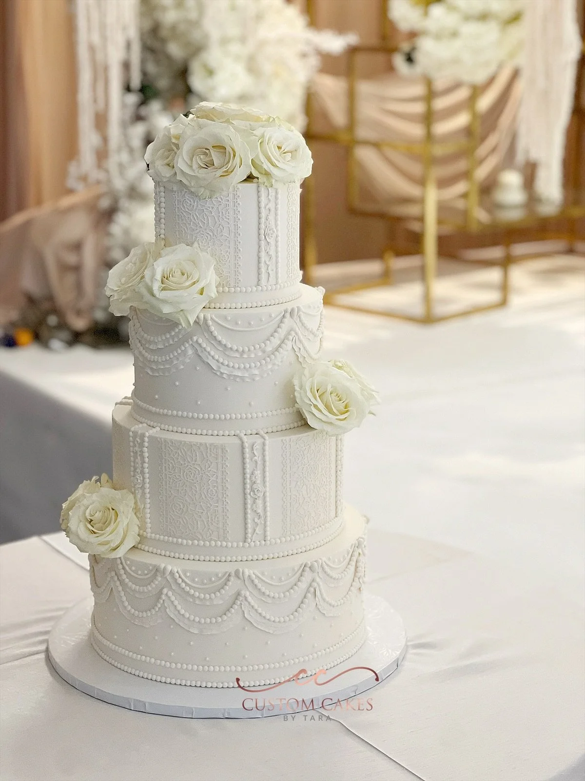 White, multi-tiered wedding cake decorated with intricate piping designs and white roses, displayed on a round silver cake board.