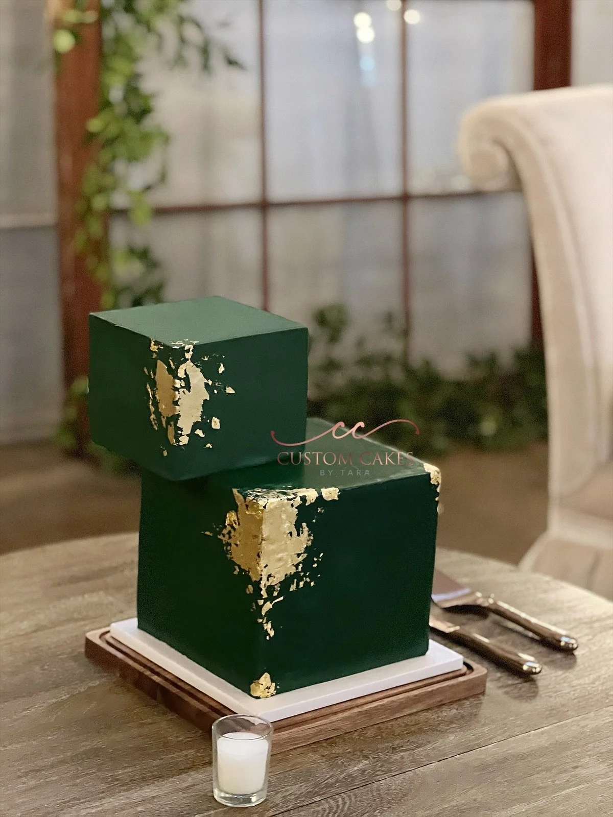 A two-tiered cake with dark green fondant and gold accents, placed on a wooden cake stand on a wooden table with a small white candle in front. There is a knife and fork beside the cake. The background features a wall with a grid pattern and some gre
