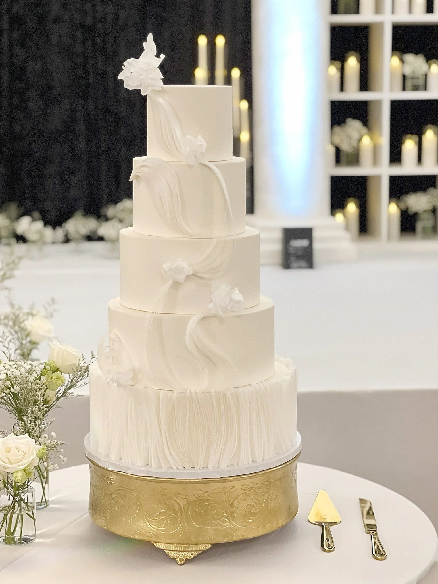 A four-tiered white wedding cake with delicate white decorative elements, placed on a gold stand. The cake is surrounded by a bouquet of white roses and green foliage in glass vases, with gold serving utensils on a white tablecloth.