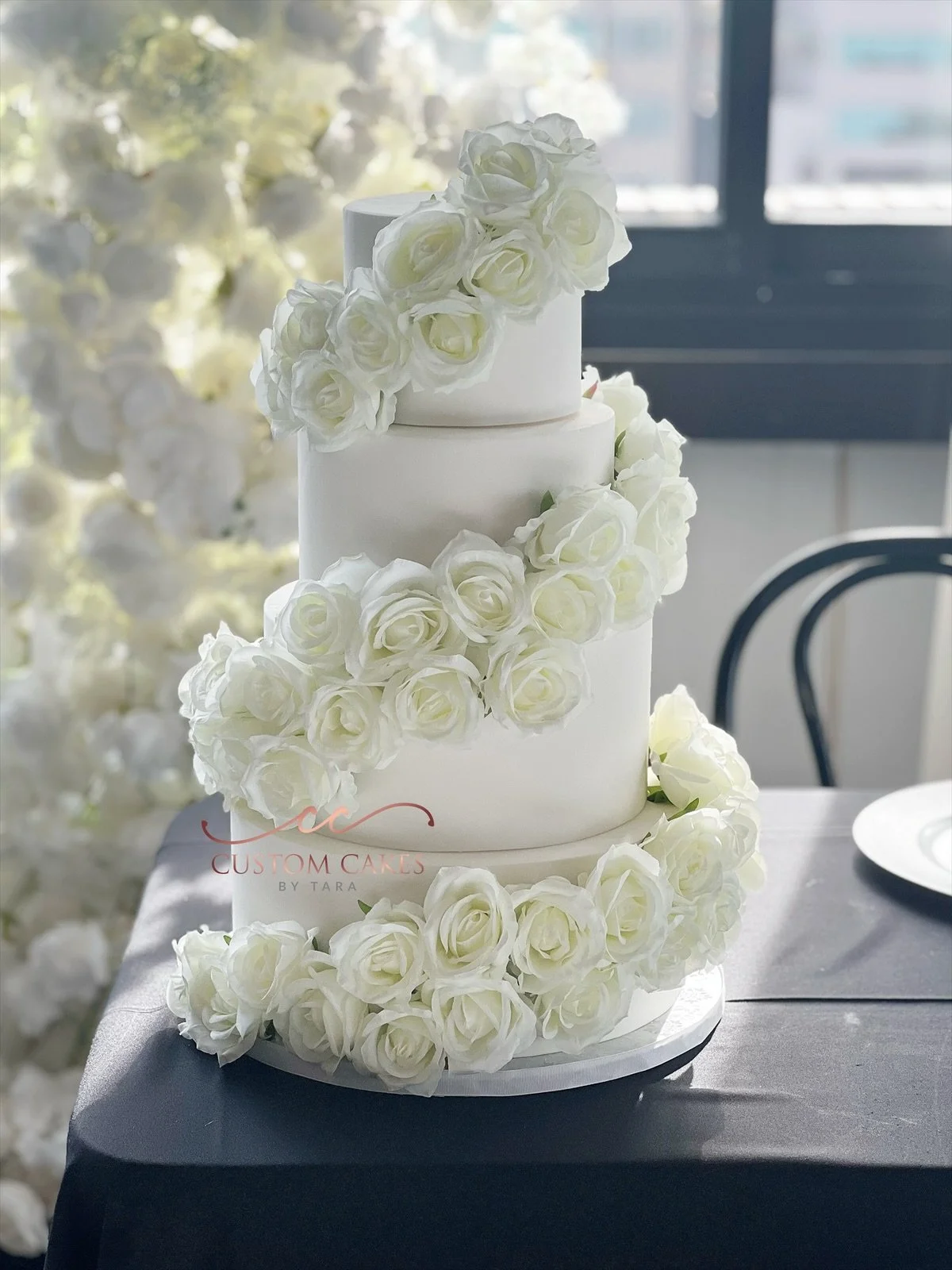 A three-tier white wedding cake decorated with white roses cascading down the side, placed on a black surface with light streaming in from windows in the background.