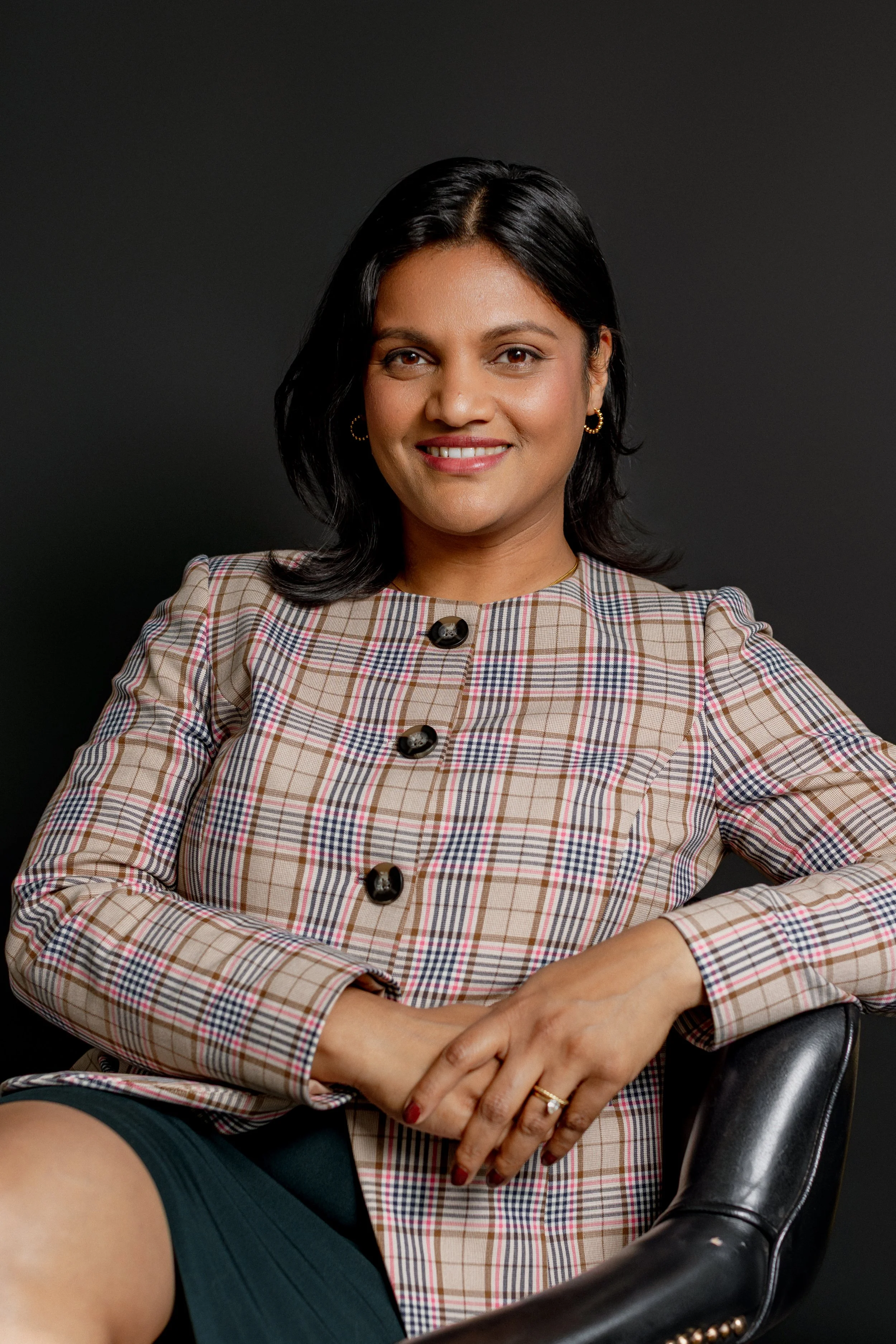 Portrait of a woman smiling, wearing a white blazer and a beige top, with dark hair and a black background.