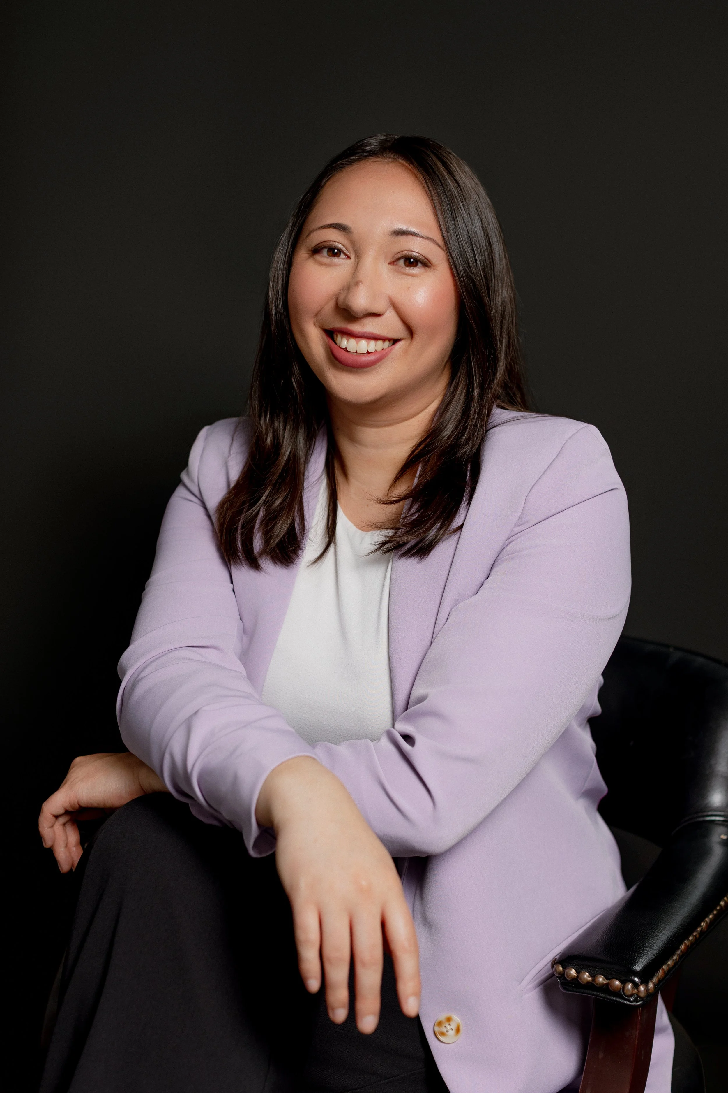 A smiling woman with long dark hair wearing a white blouse and a cream blazer, standing against a black background.